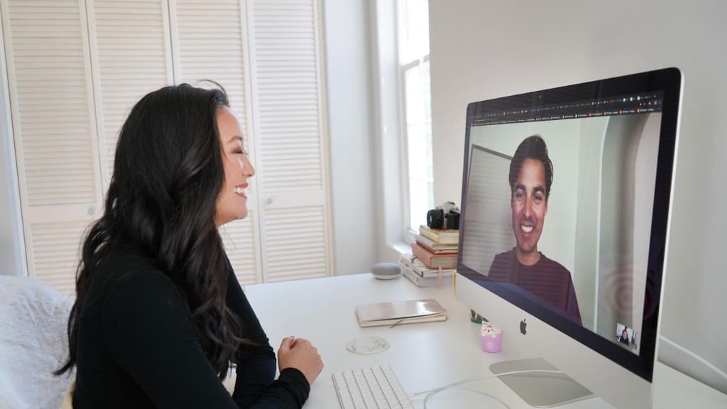 A woman sits at a desk and smiles while having a video call with a style consultant displayed on a large computer monitor in a bright, tidy room.